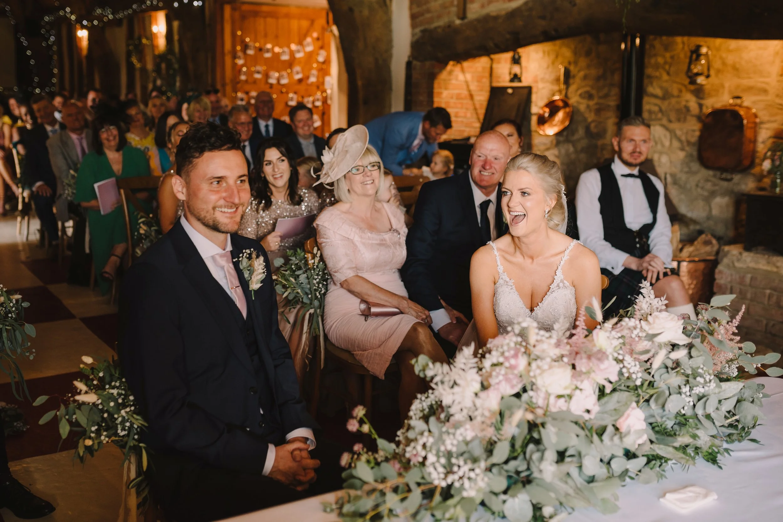 A group of wedding guests seated in a rustic venue, smiling and watching the wedding ceremony with a bride in a white wedding dress laughing and a groom in formal attire, surrounded by floral decorations.