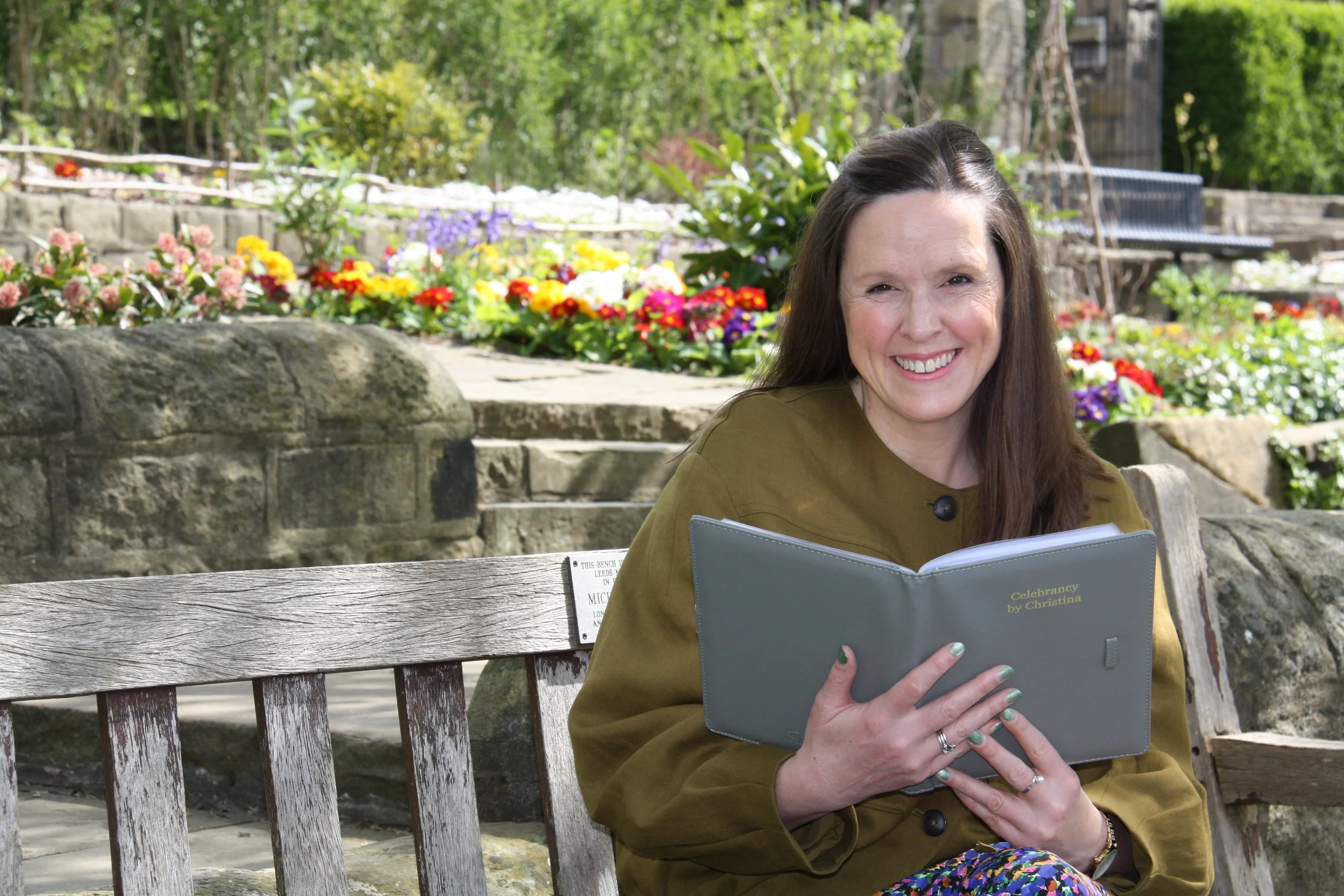 A woman with long dark hair sitting on a wooden bench in a garden setting, holding a grey folder. She is smiling and wearing an olive green jacket. In the background there are colourful flowers, trees, and a stone wall.