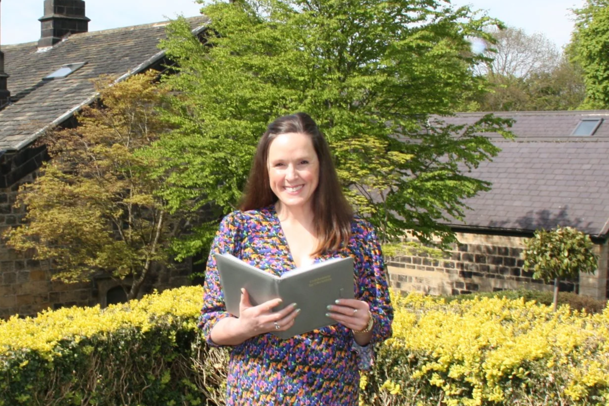 A smiling woman holding an open folder outdoors. She is standing in front of a tree with green leaves and a background of buildings and shrubs during spring.