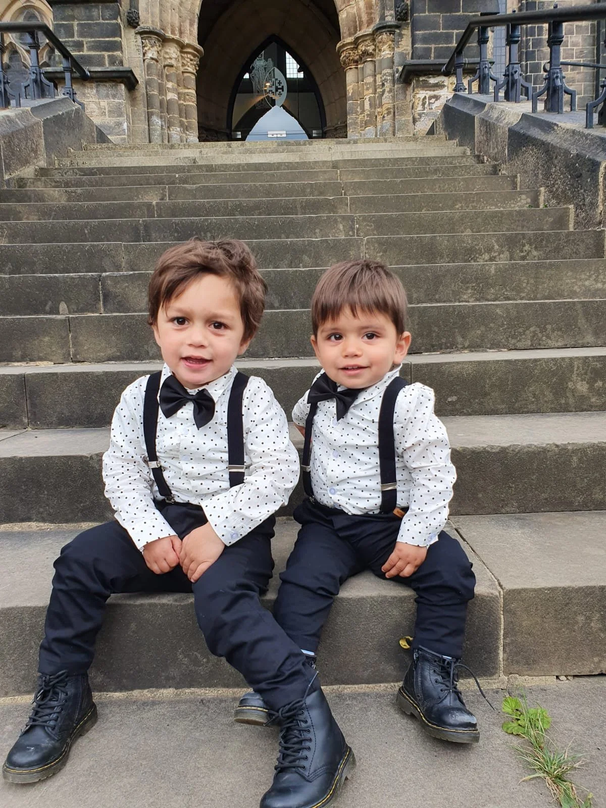 Two young boys sitting on steps in front of a historic building, wearing matching white shirts with black polka dots, black braces, black pants, black shoes, and black bow ties, posing for a photo.