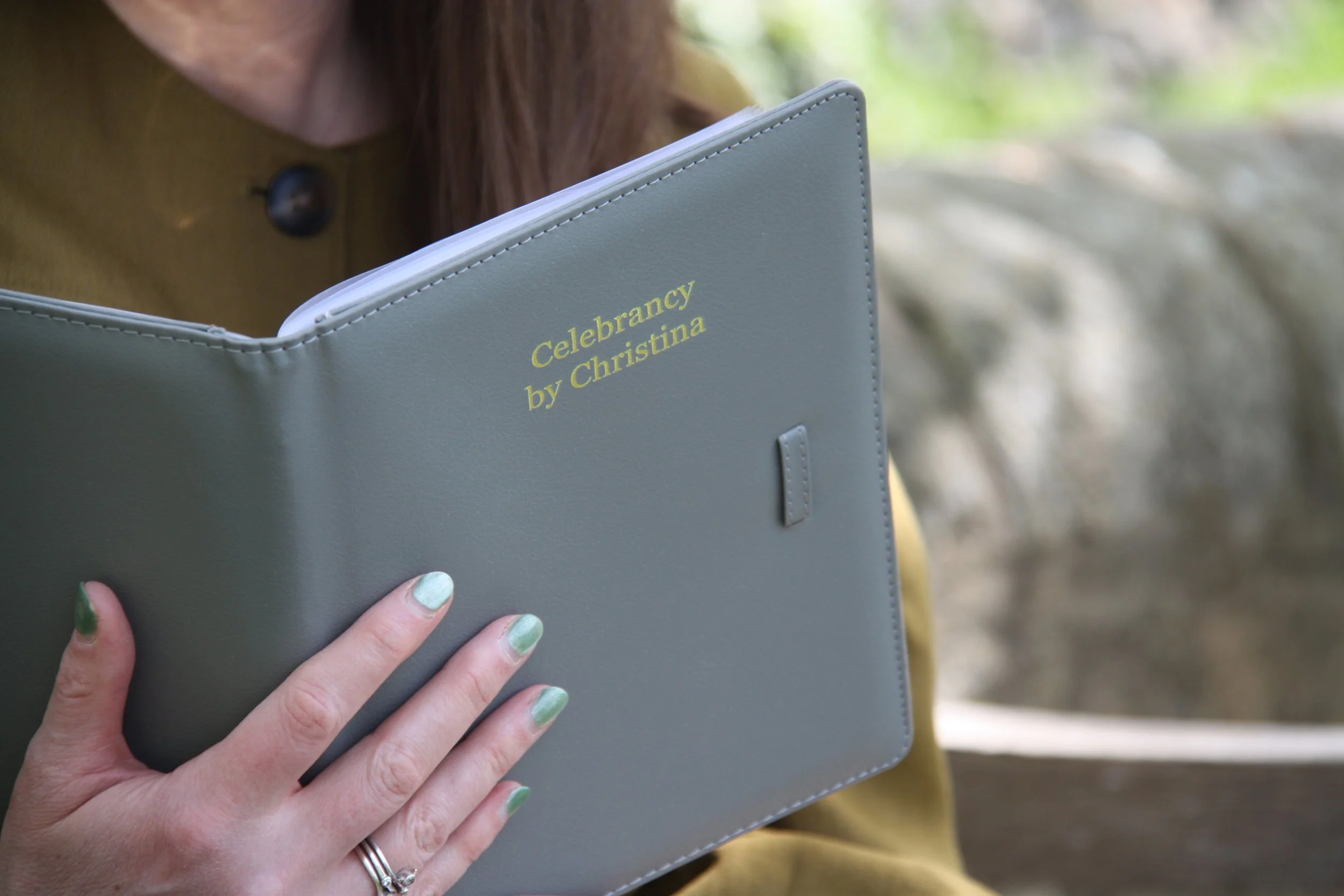 A woman holding a grey folder titled 'Celebrancy by Christina' outdoors with a blurred background.