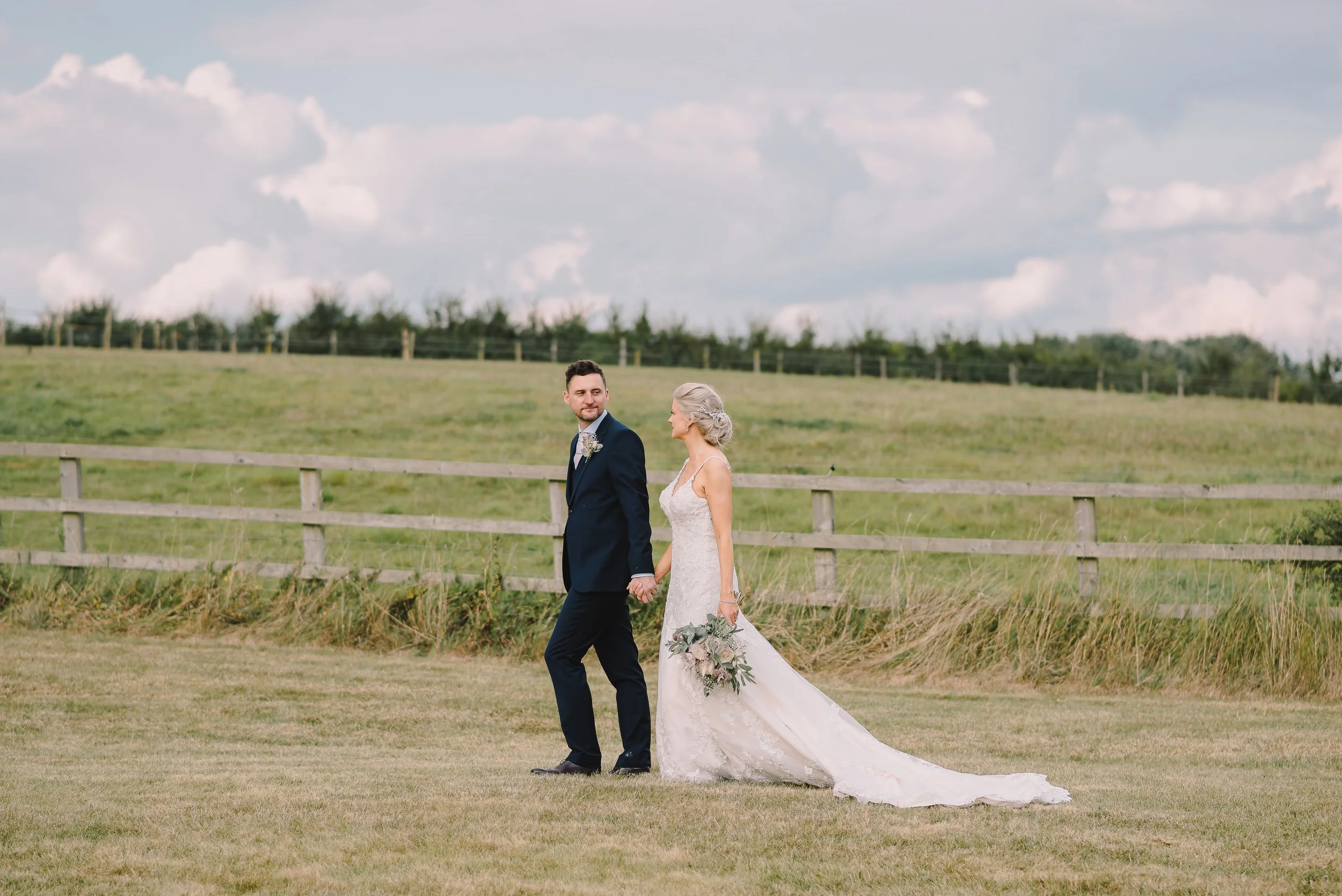 A bride and groom holding hands and walking outdoors in a grassy field, with a wooden fence and cloudy sky in the background.