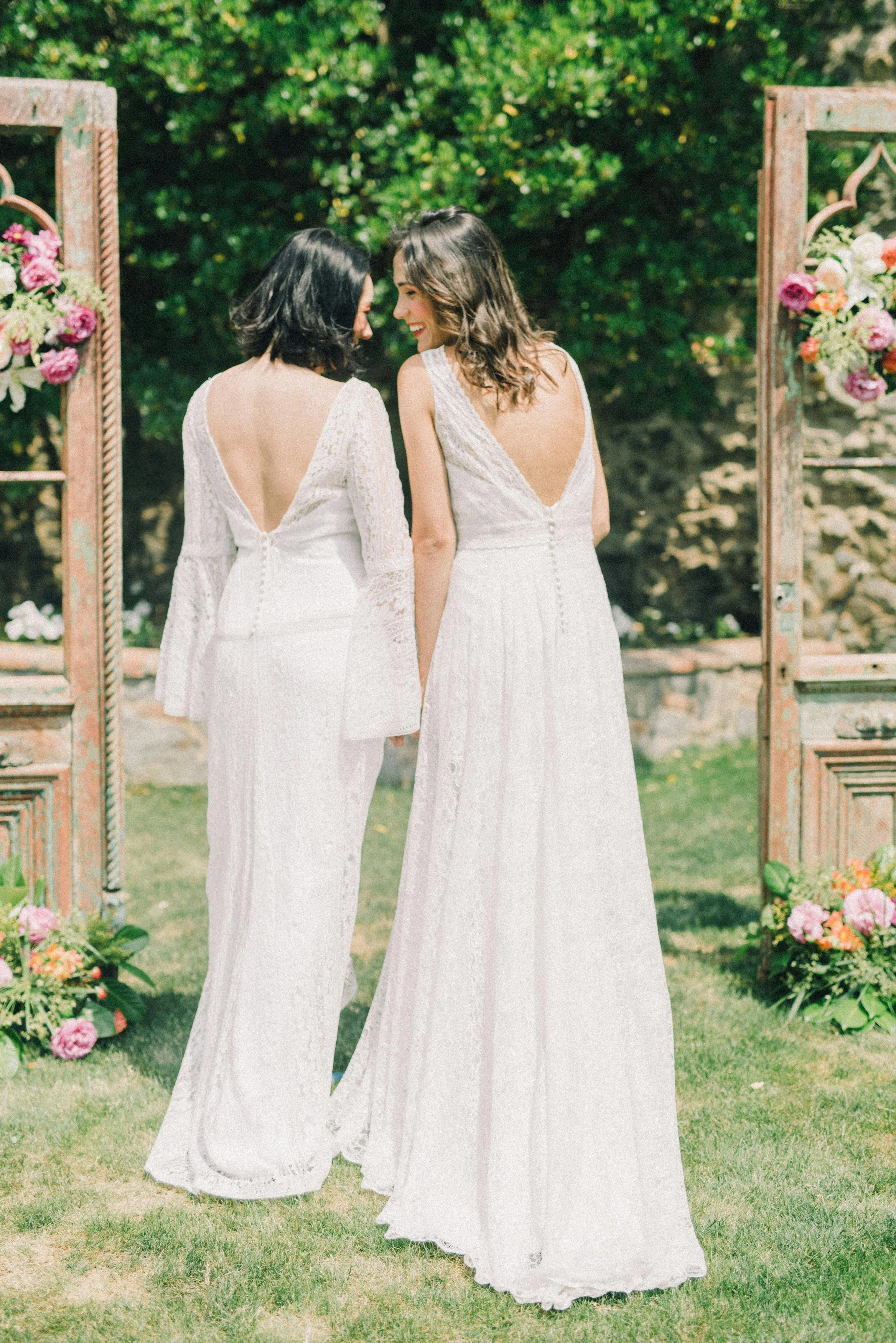 Two women in white lace dresses standing outdoors, holding hands, with their foreheads touching and smiling, at a wedding ceremony surrounded by floral decorations and wooden archways.