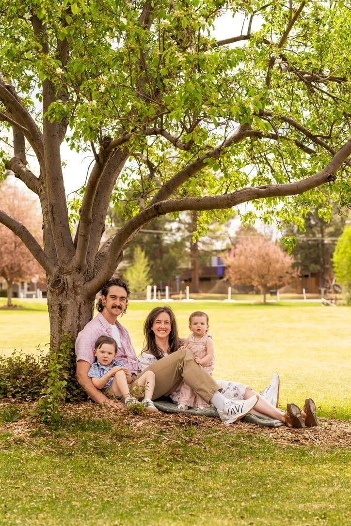 Family of four sitting and relaxing by a large tree in a park, with a grassy field and houses in the background during daytime.