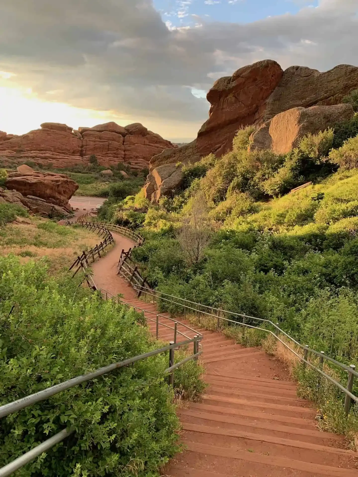 Winding dirt trail with wooden steps and metal railing surrounded by green shrubs and bushes, leading towards reddish rocks and large boulders in a desert landscape under a partly cloudy sky.
