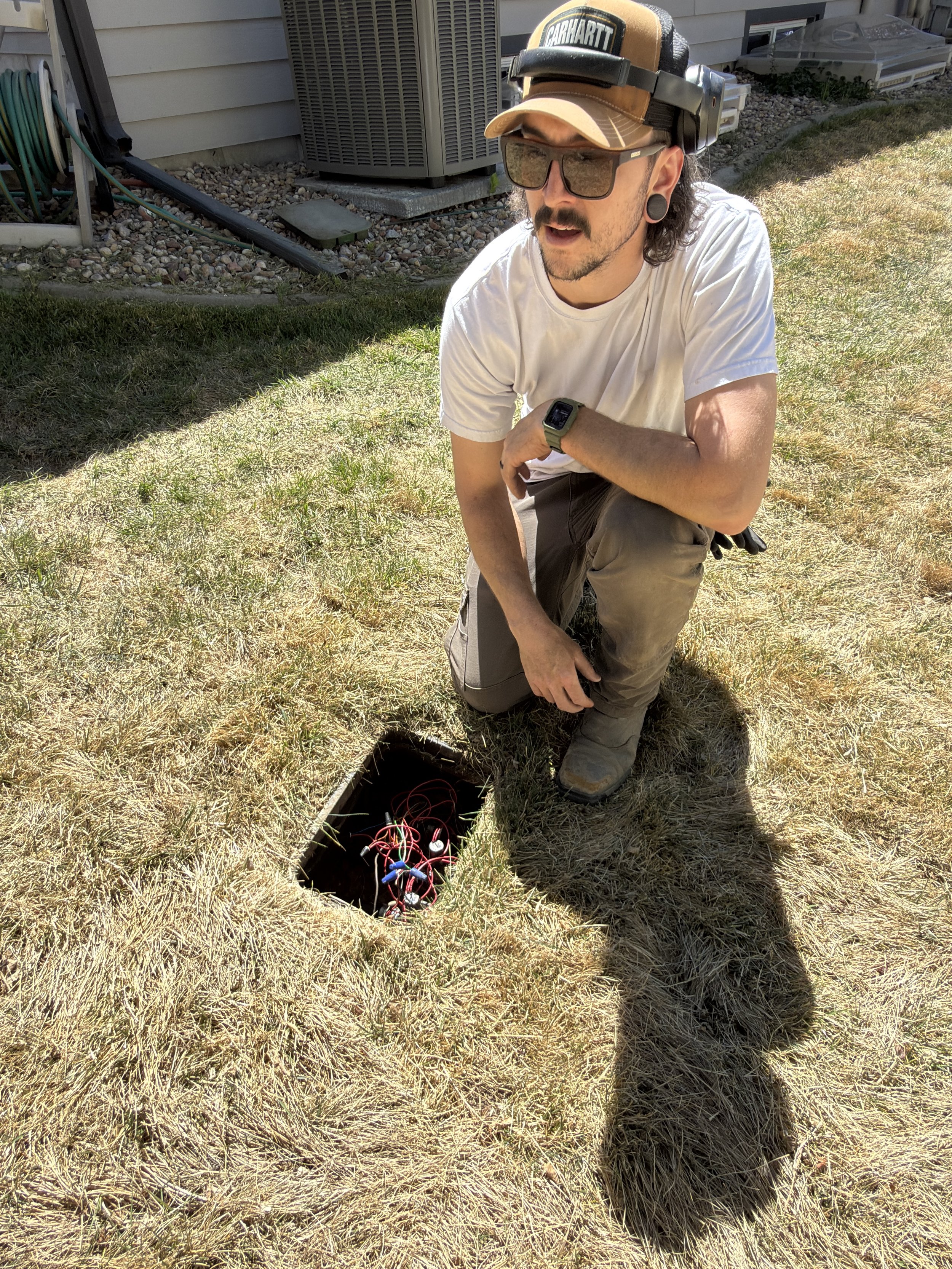 A man kneeling on a patchy grass backyard, wearing a white T-shirt, gray pants, a beige Cap with a Carhartt logo, large sunglasses, and black ear gauges. He is adjusting or inspecting the wires for an underground electrical or electrical testing device in an open hole in the ground. Behind him are an air conditioning unit, garden hose, and household items.