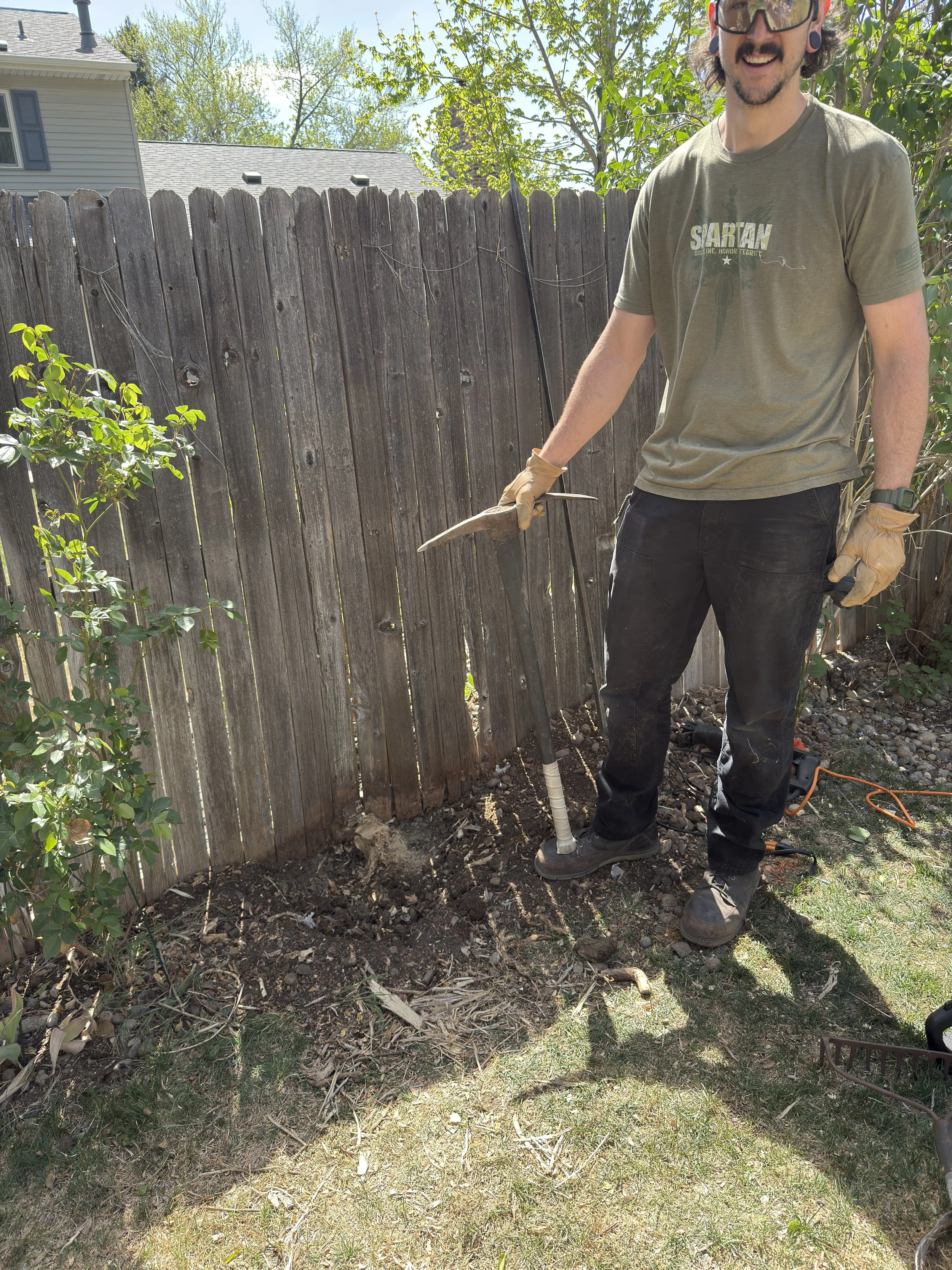 Man wearing gloves and safety glasses using a tool to dig in soil near a wooden fence in a backyard.