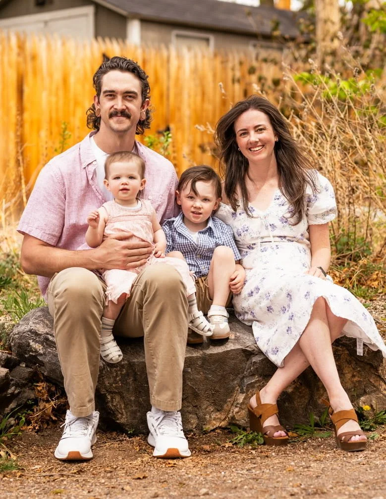 A family of four sitting outdoors on a large rock, smiling at the camera. The father has dark curly hair, a mustache, and is wearing a pink shirt and khaki pants. The mother has long brown hair and is wearing a white dress with purple floral patterns. The young girl, sitting on the father's lap, has short hair and is wearing a light pink dress. The young boy sitting next to the mother has short brown hair, a checkered shirt, and shorts. The background features a wooden fence, dry plants, and a house.