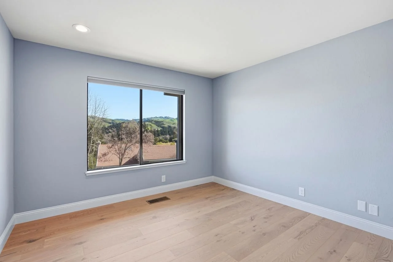 Empty room with light blue walls, a window showing a view of trees and hills, hardwood floors, and white baseboards.