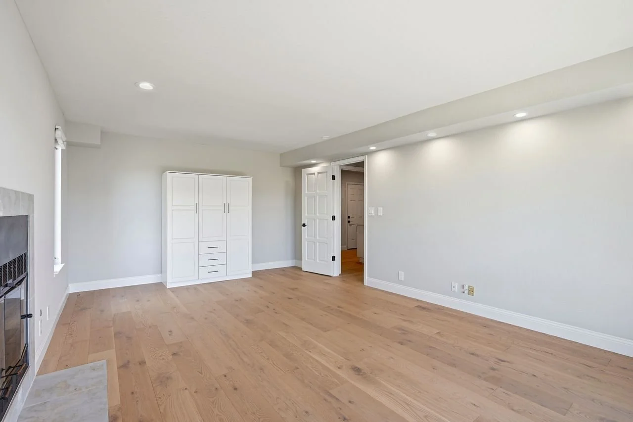 Empty living room with light wooden floors, white walls, recessed ceiling lights, a white cabinet, a fireplace, and an open door leading to another room.