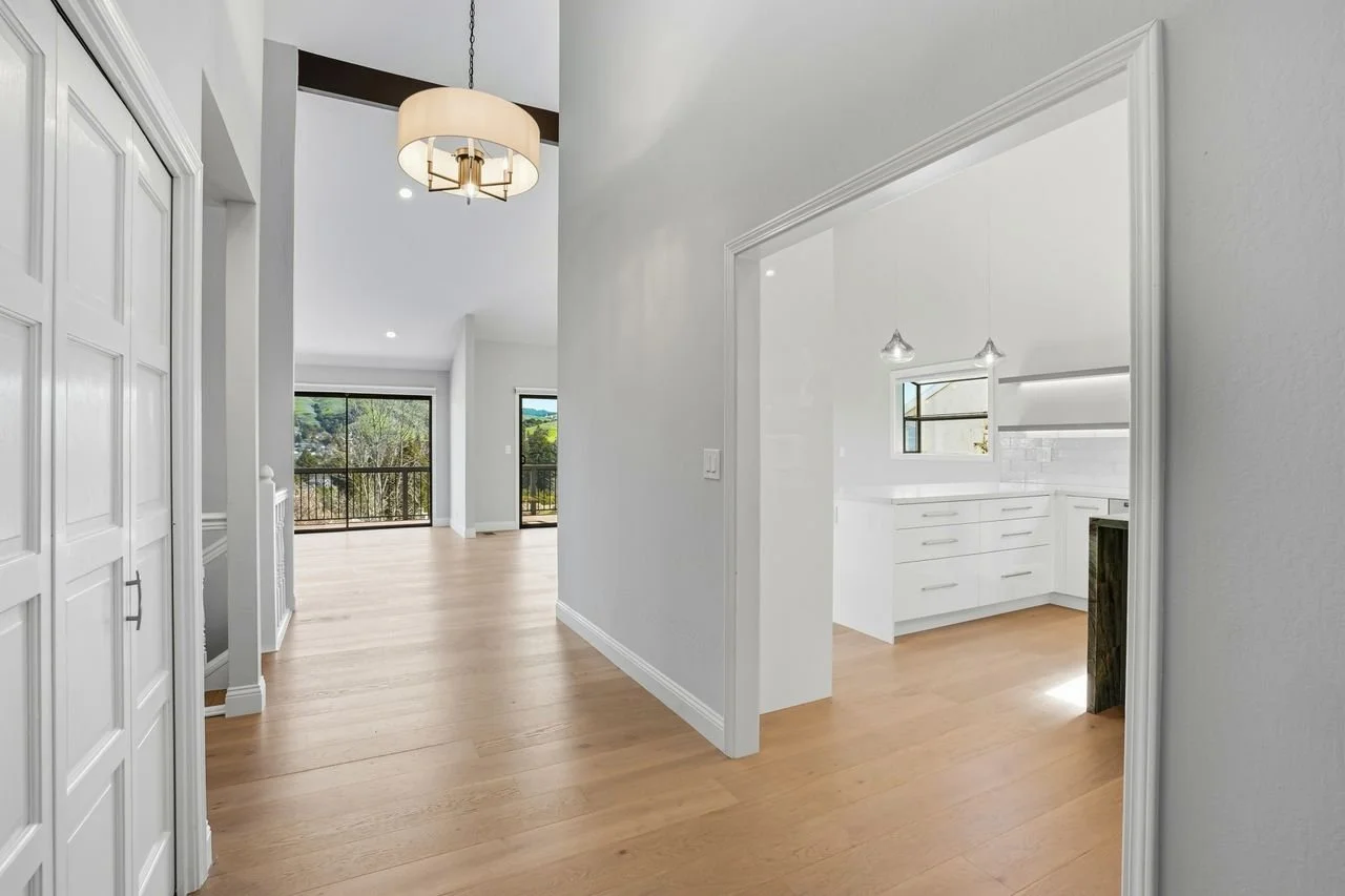 Bright, open living space with large windows, light wood flooring, and white walls. An area with a kitchen visible through an opening, featuring white cabinetry and a window. A sliding glass door leading to a balcony or patio area.