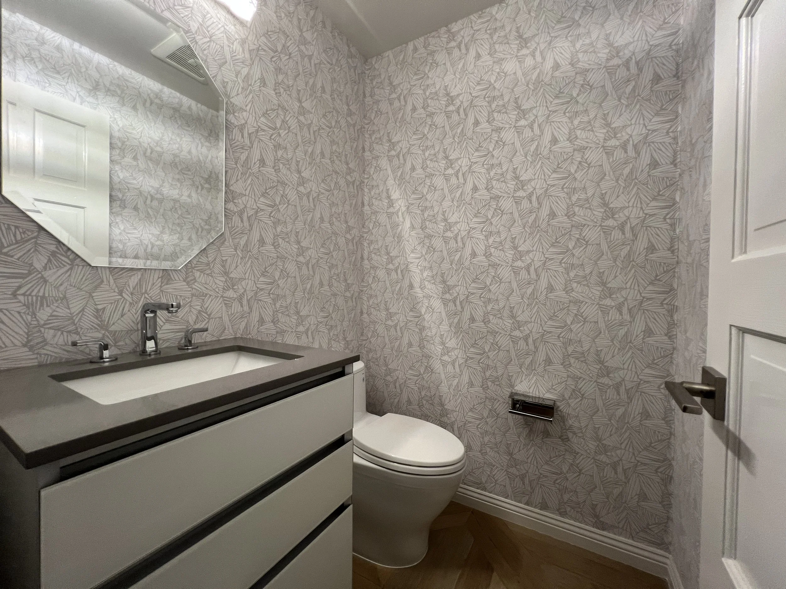Small bathroom with a geometric patterned wallpaper, a white vanity with a sink and silver faucet, a wall-mounted mirror, a toilet, a toilet paper holder, and a partially open door.