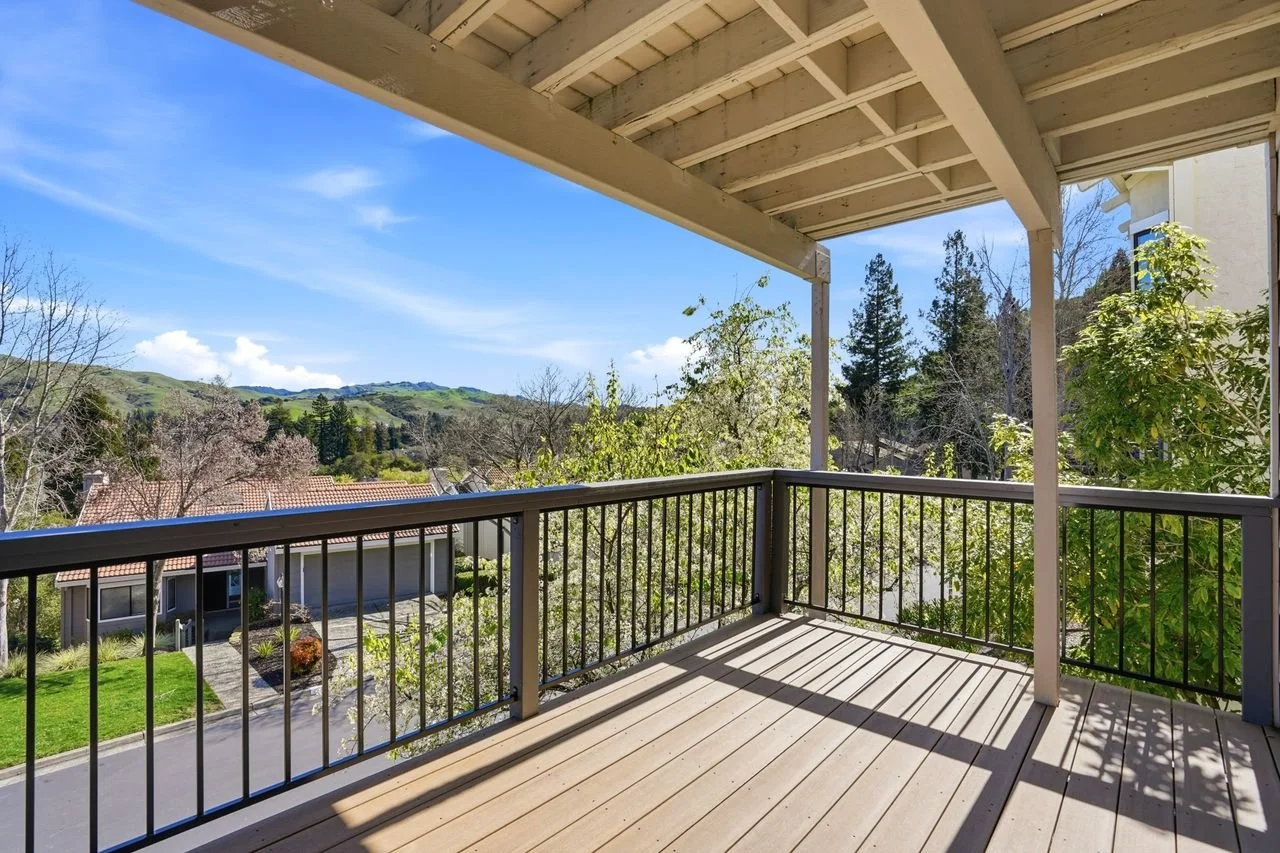 View from a balcony with wooden flooring and black metal railing overlooking a suburban neighborhood with single-story houses, trees, and hills under a blue sky with some clouds.