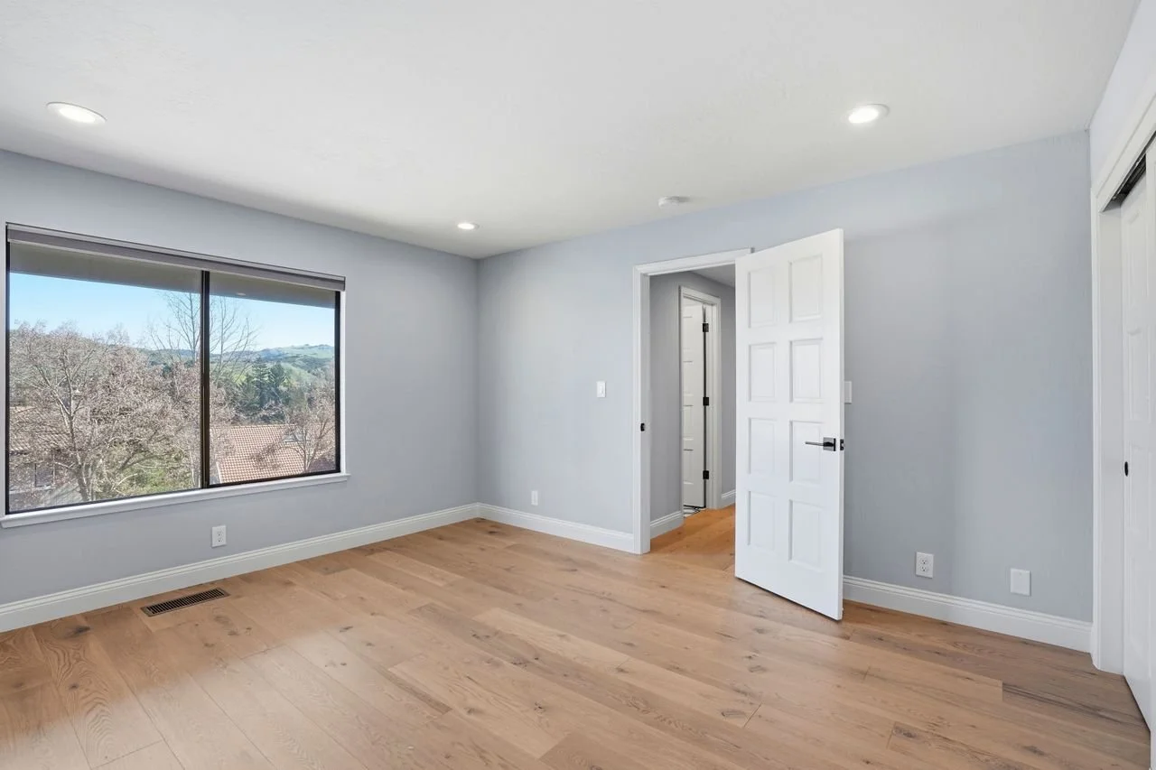 Empty bedroom with large window, light blue walls, white door, hardwood floor, and ceiling lights.