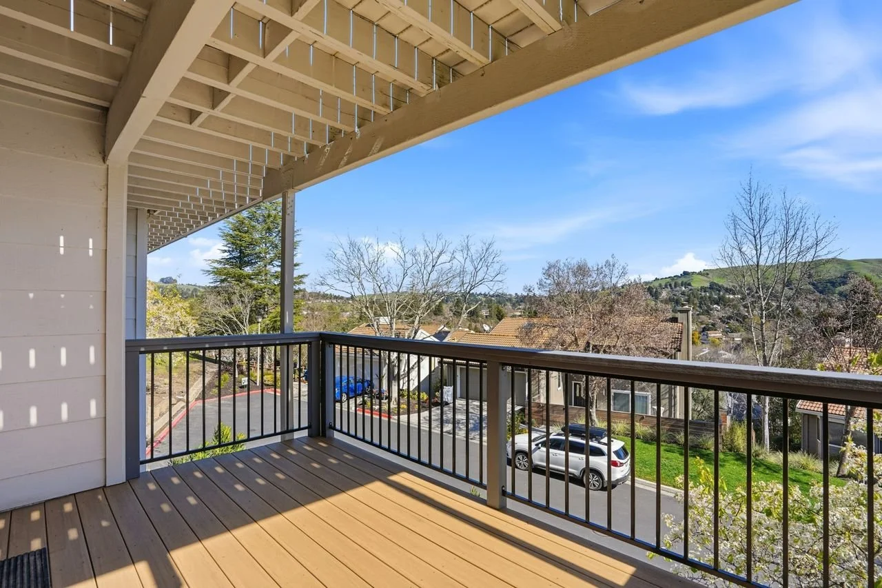View from a balcony with black railing, showing houses, trees, and distant green hills under a blue sky with wispy clouds.