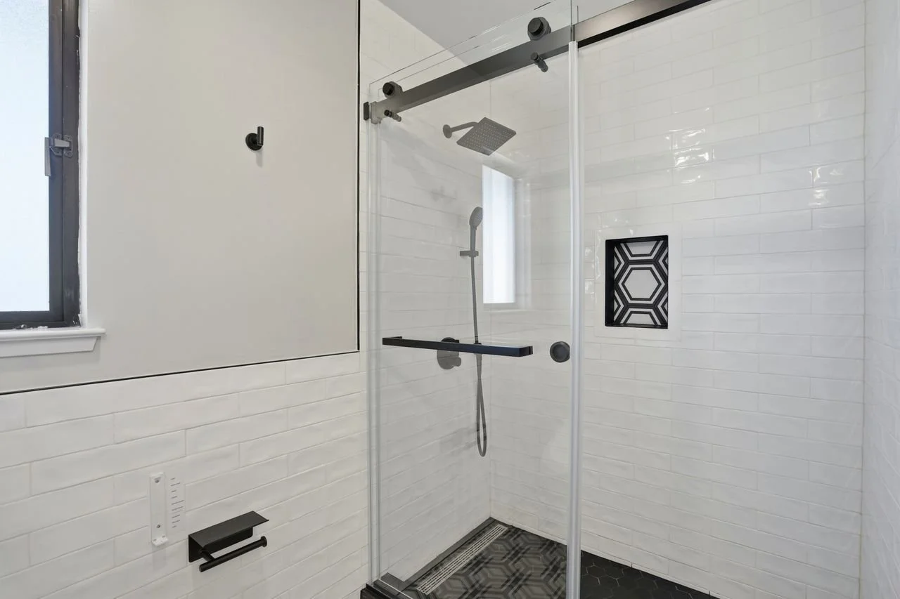 Modern bathroom with a glass shower enclosure, black fixtures, white subway tile walls, black hex tile floor, and a small window.