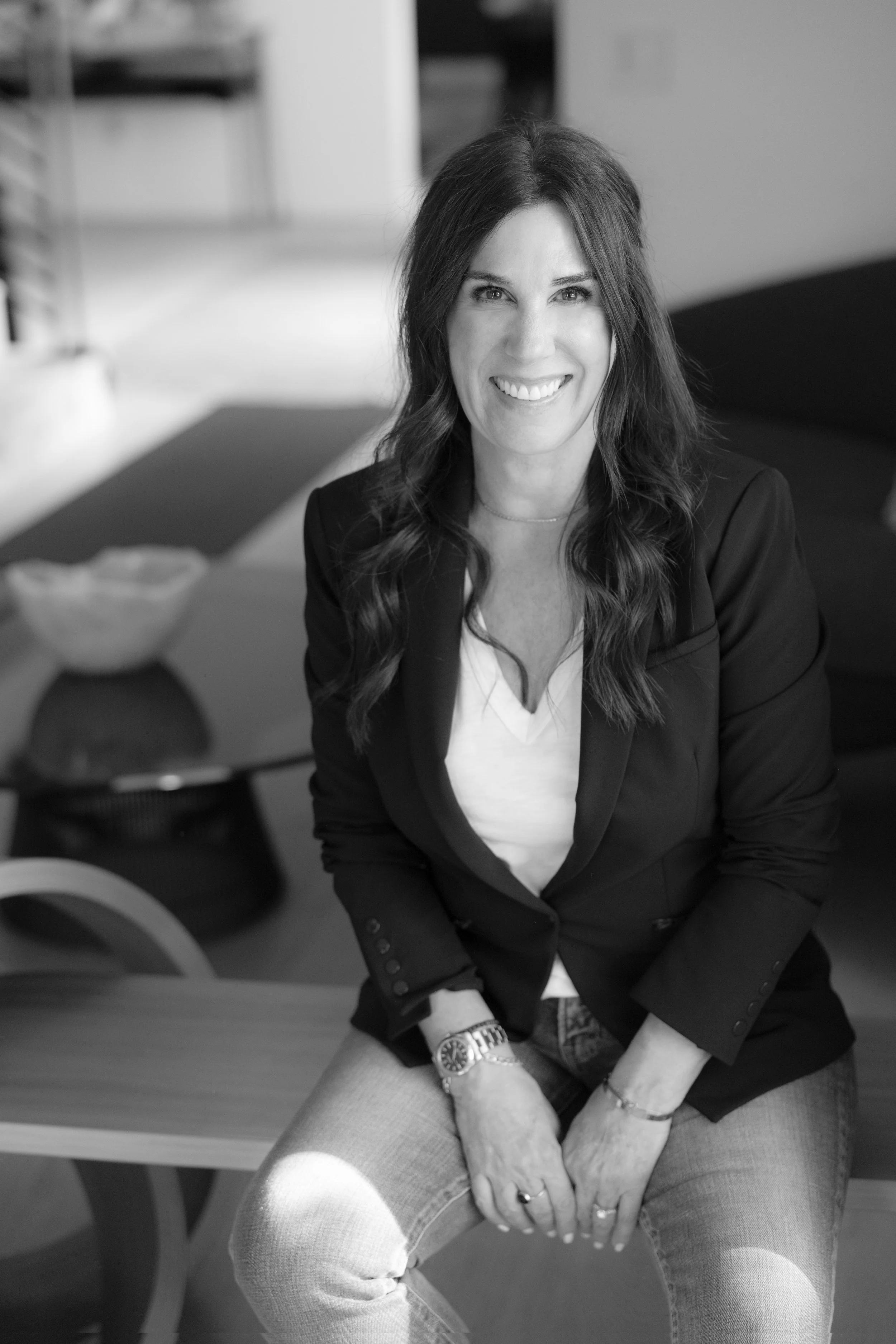A woman with long dark hair is smiling and sitting on a bench in an indoor setting.