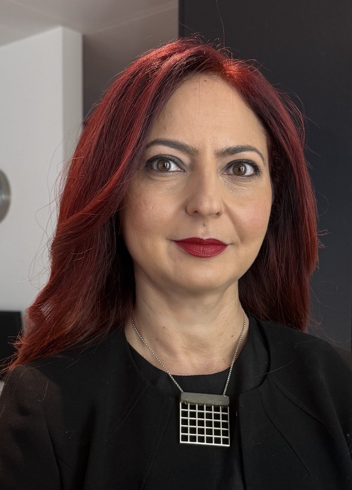 A woman with red hair and brown eyes wearing a black top and a geometric silver necklace.