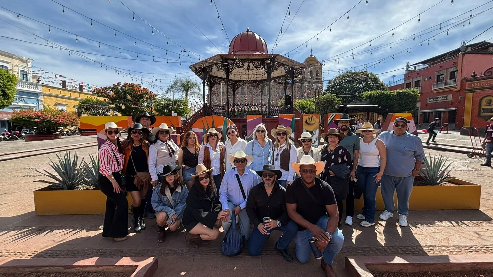 Grupo de personas en una plaza con un quiosco decorativo y luces, vistiendo sombreros y gafas, posando para la fotografía.