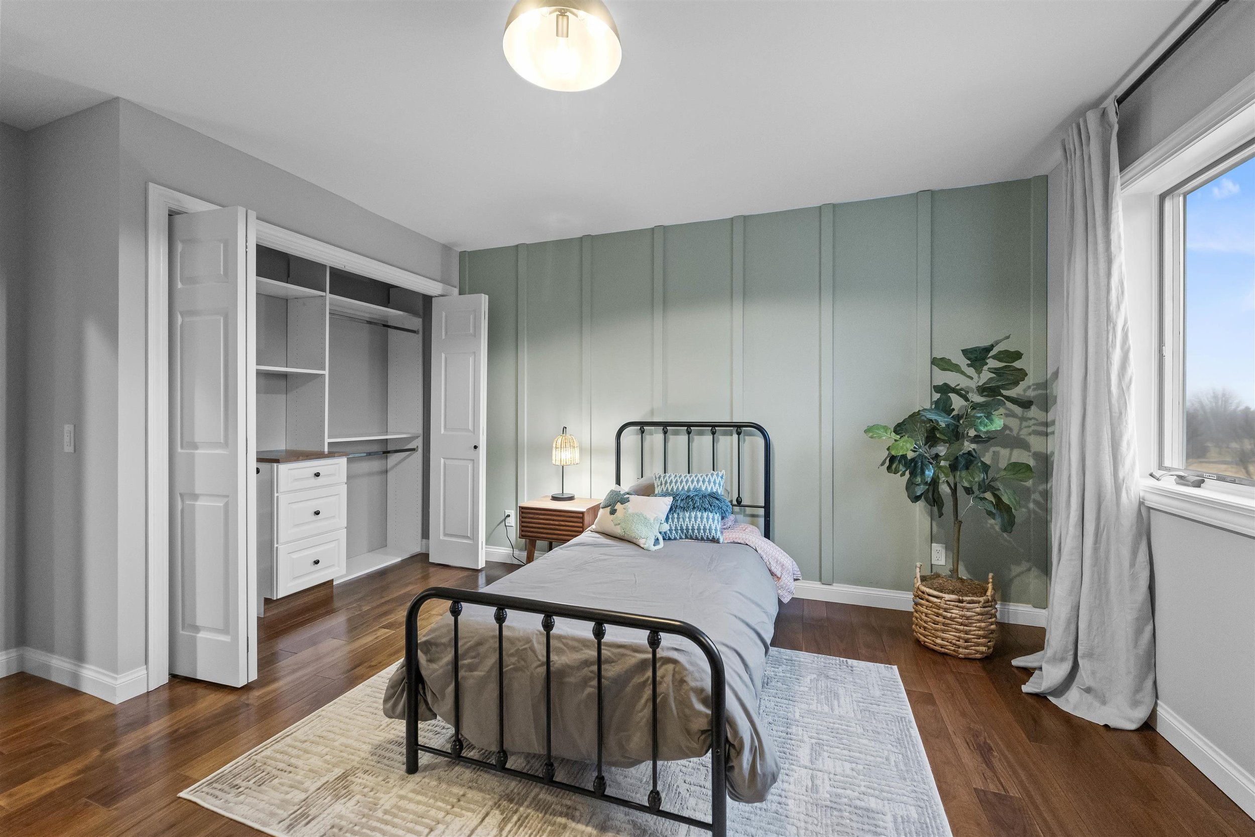 A bedroom with a metal twin bed, a nightstand with a lamp, and a large window with curtains. There is a potted plant in a wicker basket and an open closet with shelves and drawers. The room has hardwood floors and a green accent wall.