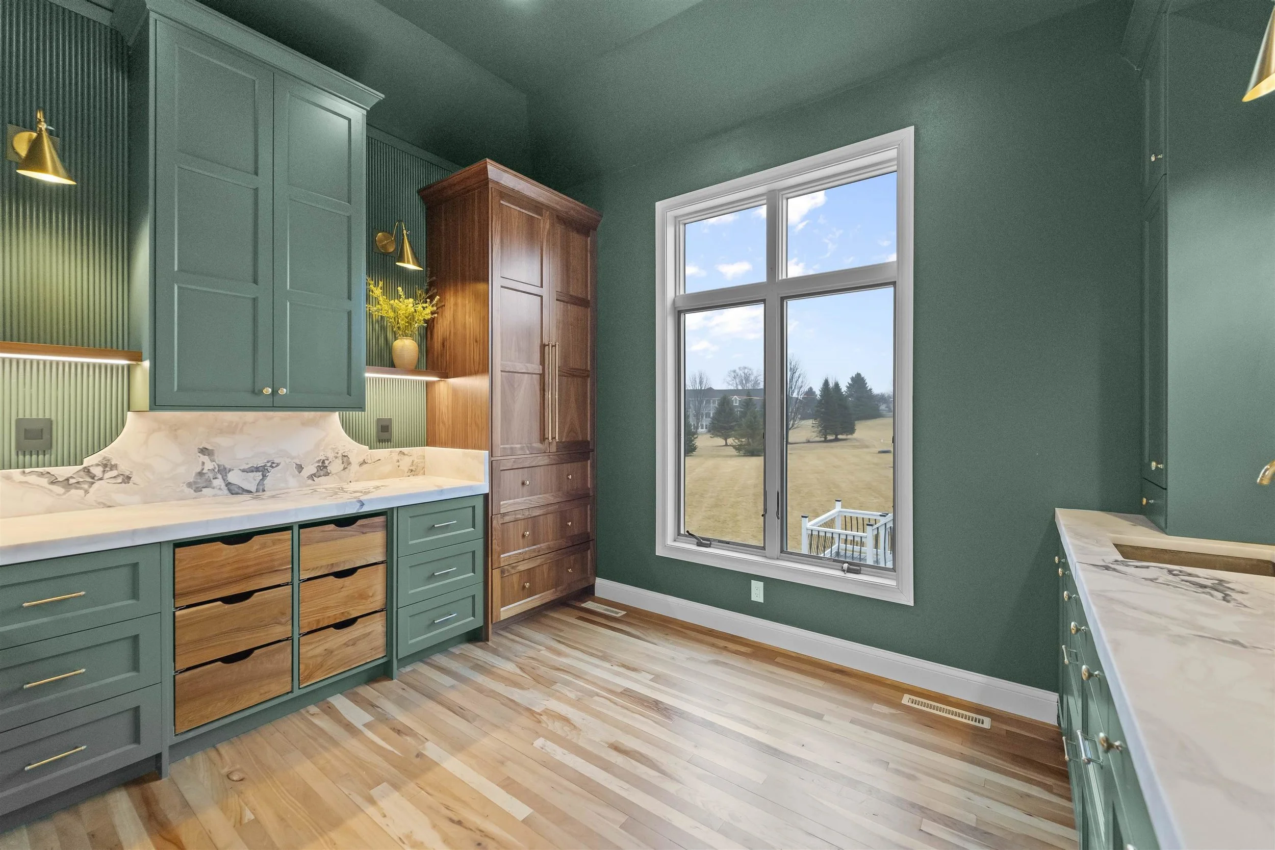 Kitchen with green cabinets, white marble countertops, a large window overlooking a backyard, and wooden flooring.
