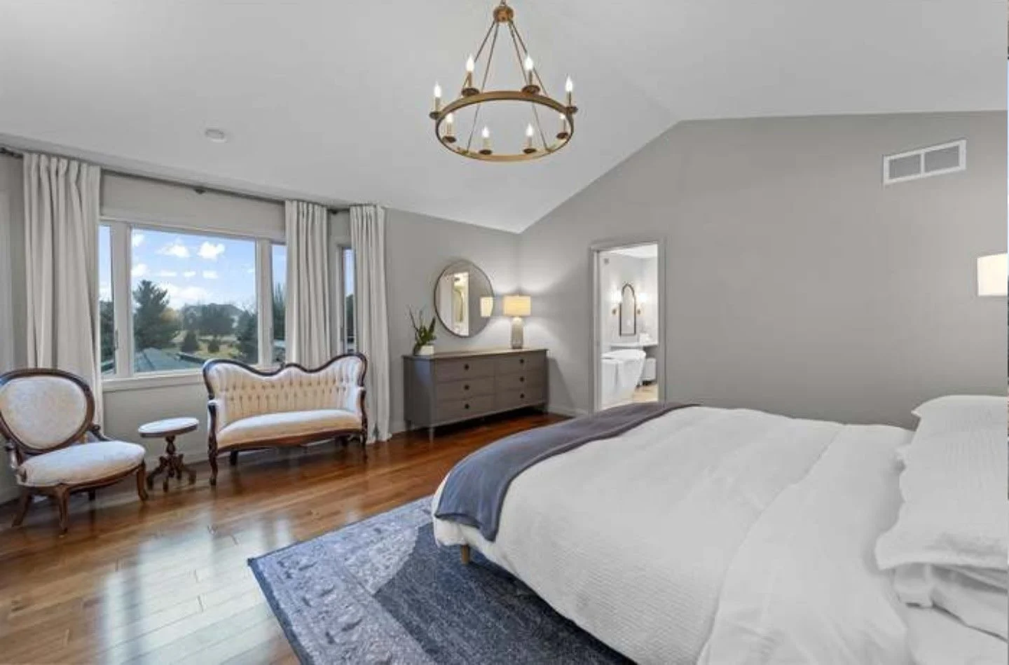 A bedroom with a large bed, a gray dresser, a mirror, a chandelier, and a sitting area near a window with white curtains, overlooking a view of trees and sky.