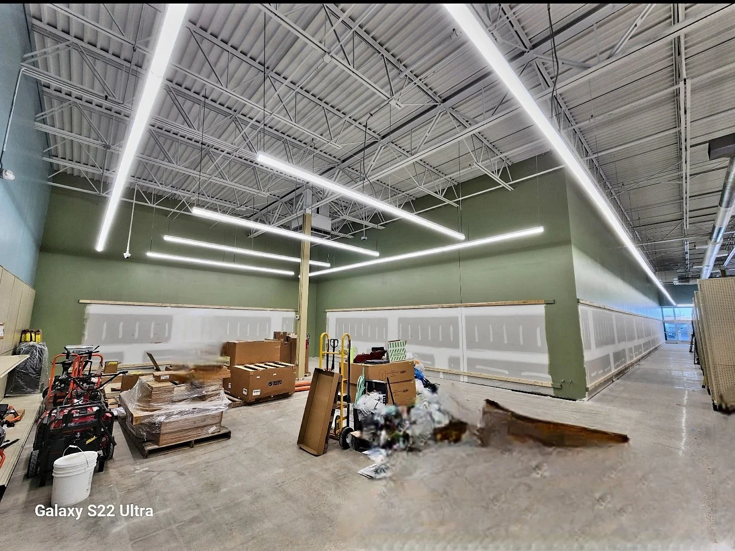 Interior of a retail store under construction, showing green painted walls, unfinished white drywall, exposed ceiling with metal beams and lighting fixtures, and scattered construction materials and tools on the floor.