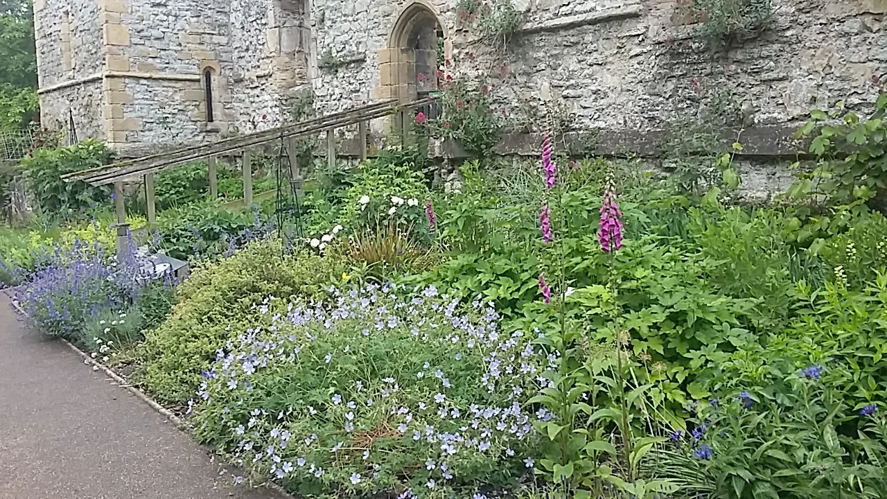A lush garden Archbishop's Palace with green plants and colourful spring summer flowers next to a stone building or castle wall.
