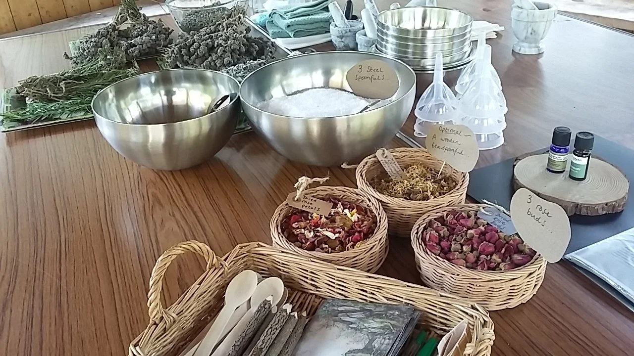 A wooden table holds two large metal bowls filled with herbs, with small handwritten labels. Wicker baskets contain dried flowers and petals. Bottles of essential oils, a stack of metal bowls, scoops and other botanical items pencils parchment paper