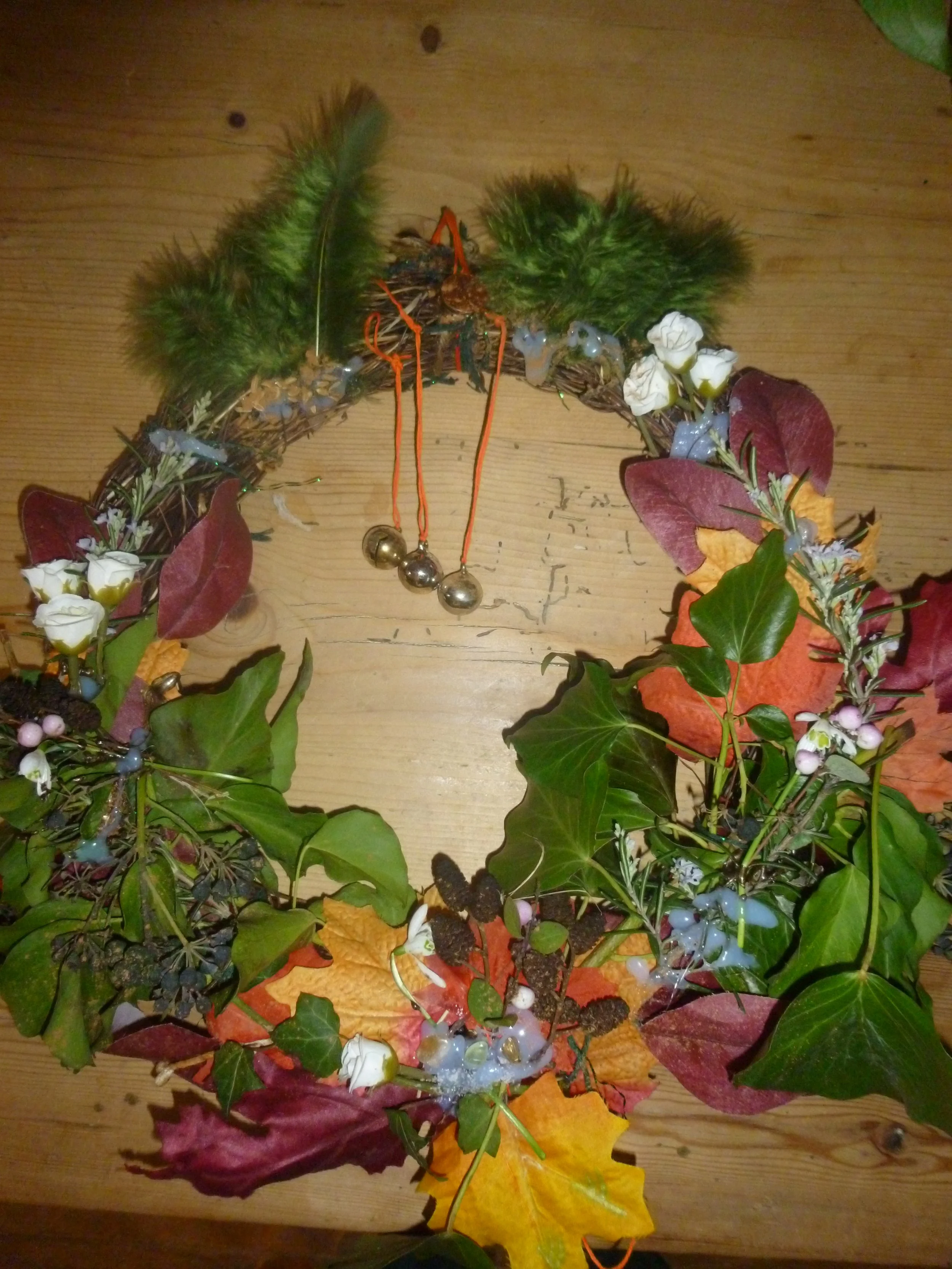 A handmade autumn and winter solstice Yule Christmas wreath made of various colourful leaves, berries, pinecones, white flowers, bells and green feathers, spell and intention intent prayer hanging on a wooden surface.