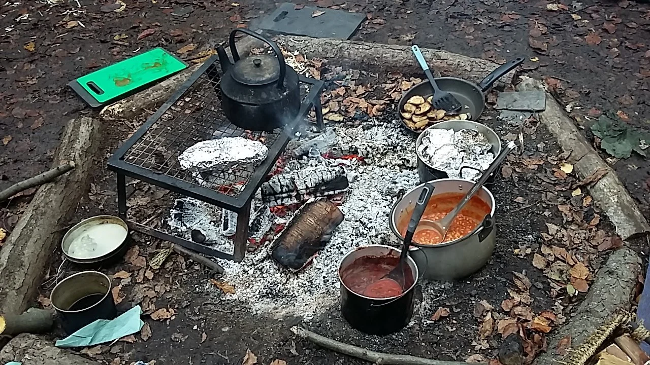 Camping cooking setup with camp fire bonfire pots, pans and food on a campfire surrounded by a ring of logs in a forest. Log circle. Forest school cooking.