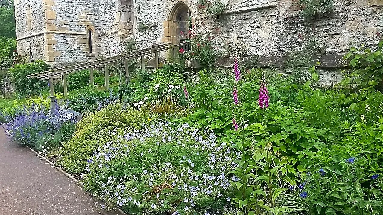 A lush garden with various colorful flowers growing along a stone wall, next to a castle or historical building with an arched doorway and small windows.