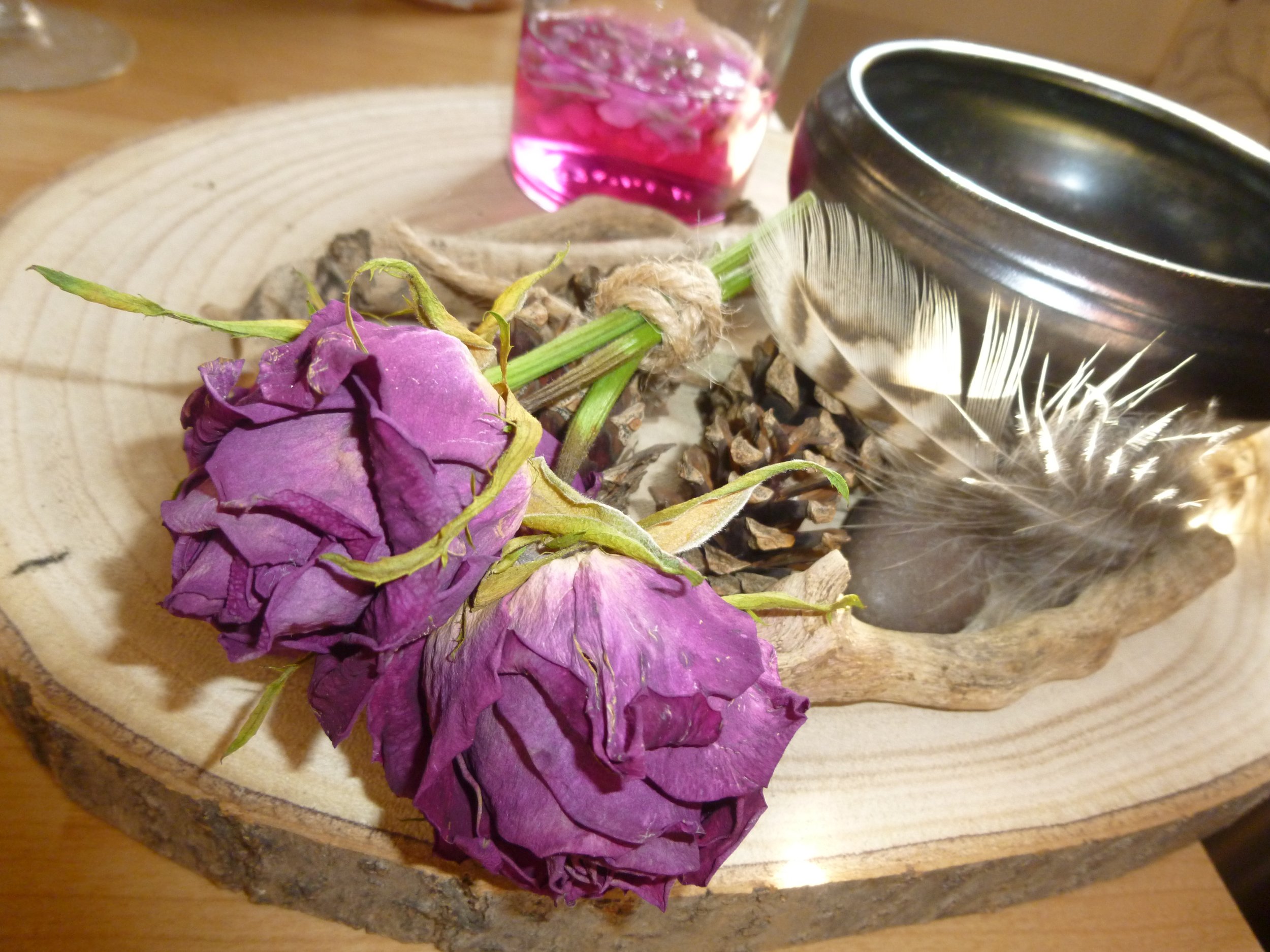 Real Dried purple roses and pinecones on a wooden tree circle slab with a pink violet potion elixir and a feather.