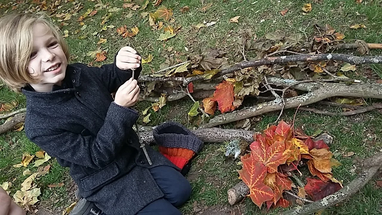 A handsome young boy with blond hair sitting on grass, smiling, surrounded by fallen autumn leaves and sticks he gathered for a craft project. Making fire. Forest school art craft mandala nature based learning play creative.