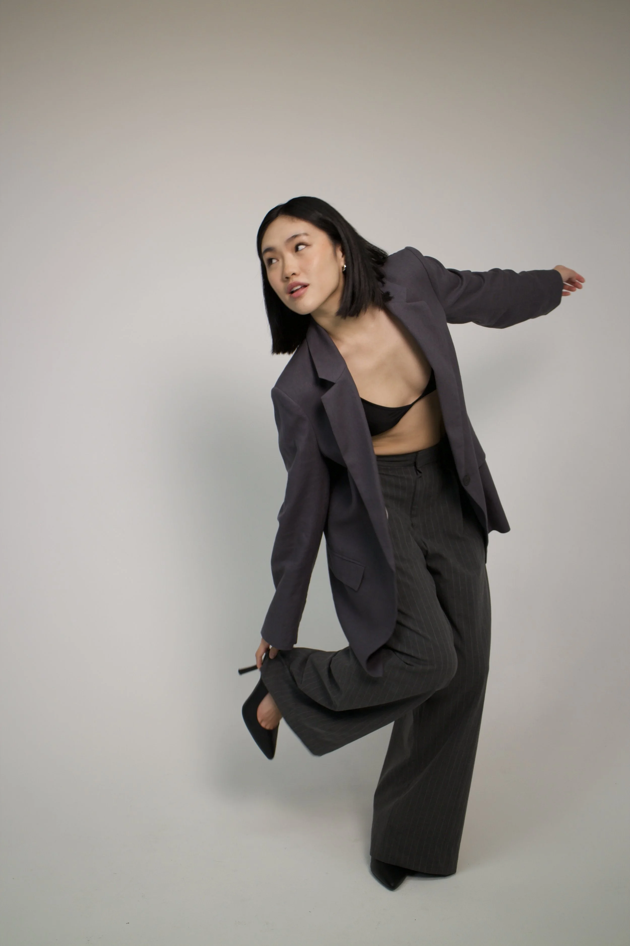 A woman with black hair, wearing a gray blazer and pinstripe pants, balancing on one leg while holding a black high heel shoe.