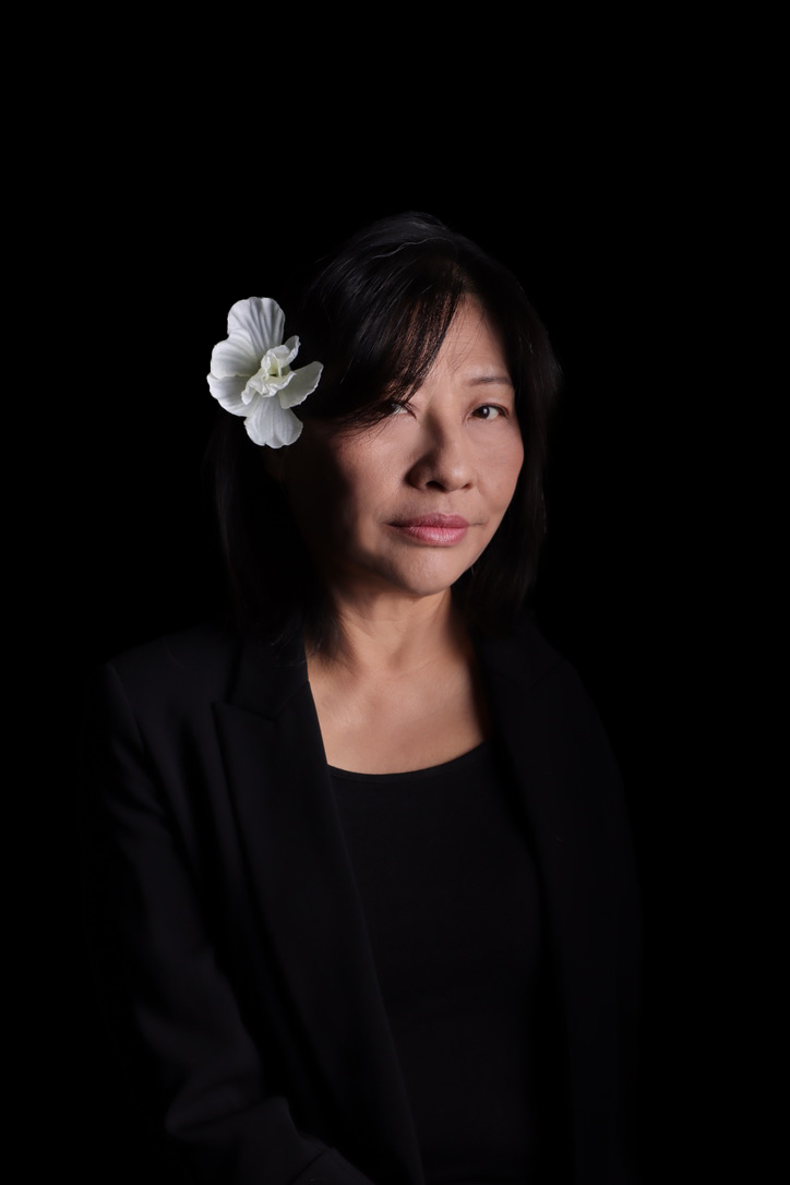 A woman with black hair and a white flower in her hair, wearing a black blazer and top, looking at the camera against a black background.