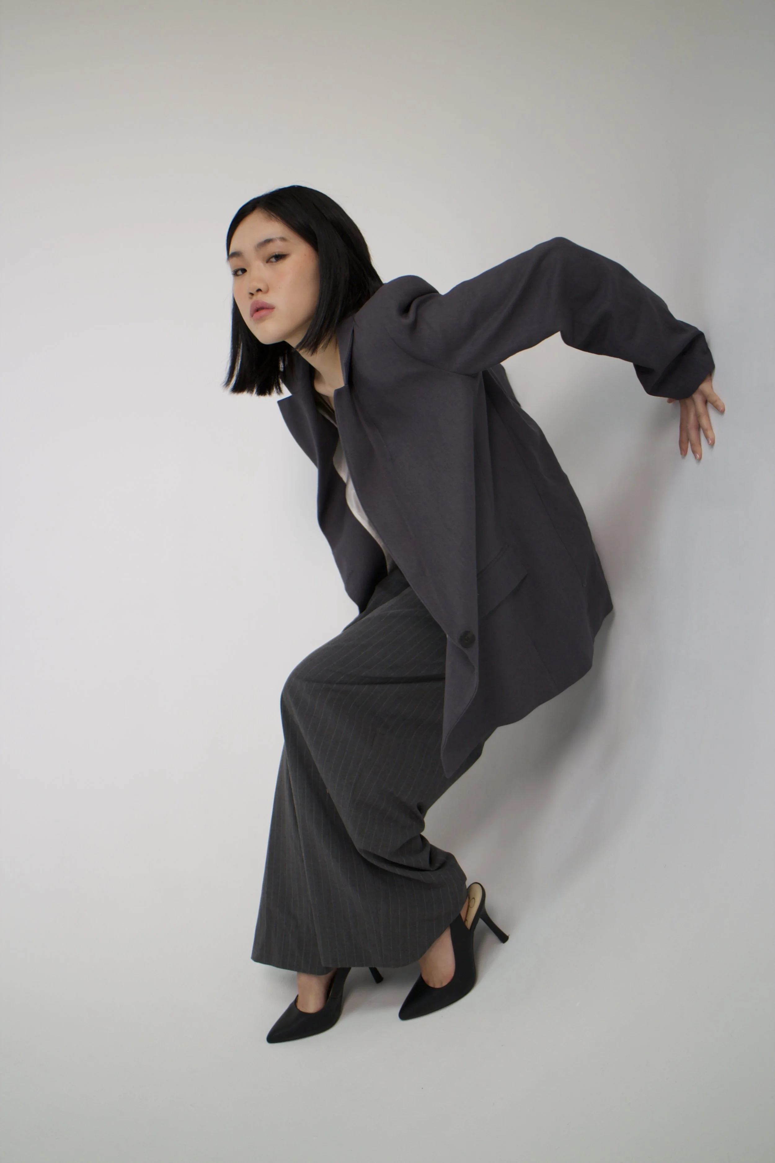 A woman with short black hair in a pose leaning against a light-colored wall, wearing a dark blazer, pinstriped trousers, and black high heels.