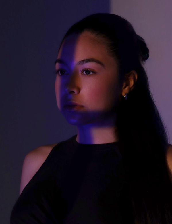 Portrait of a young woman with long dark hair, wearing a black sleeveless top, illuminated by purple and blue lighting, against a plain background.