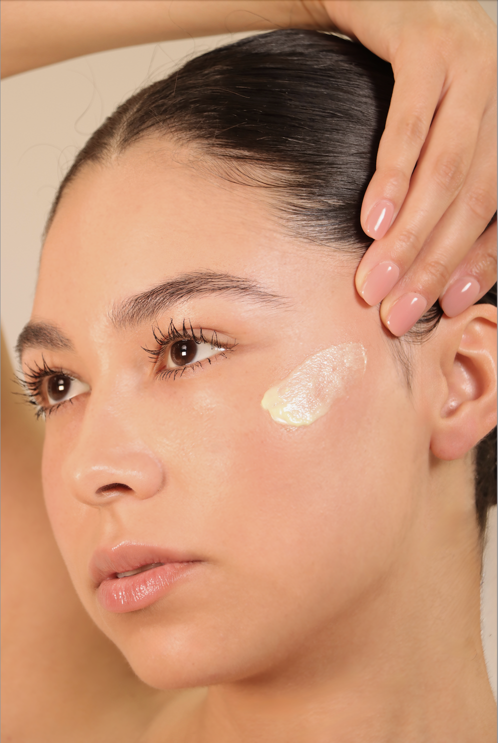 Close-up of a woman applying moisturizer to her cheek, with part of her face and hand visible, and her dark hair pulled back.