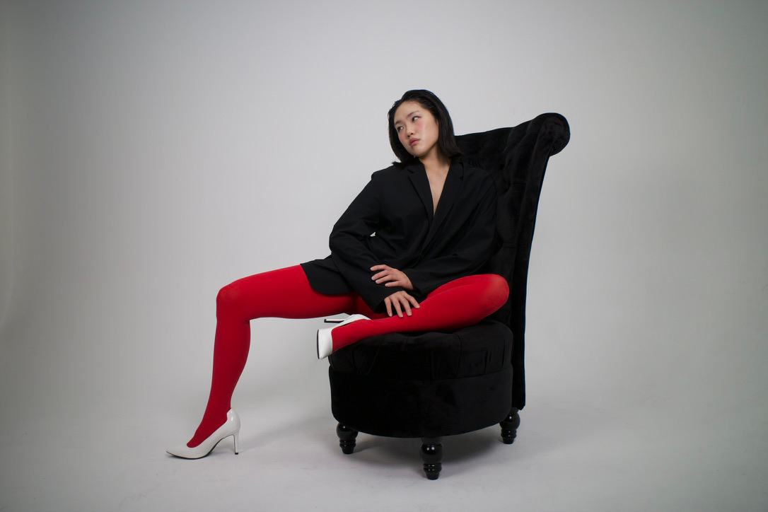 A woman with dark hair, wearing a black blazer, red tights, and white high heels, sits on a black armchair against a plain white background, looking thoughtfully to the side.