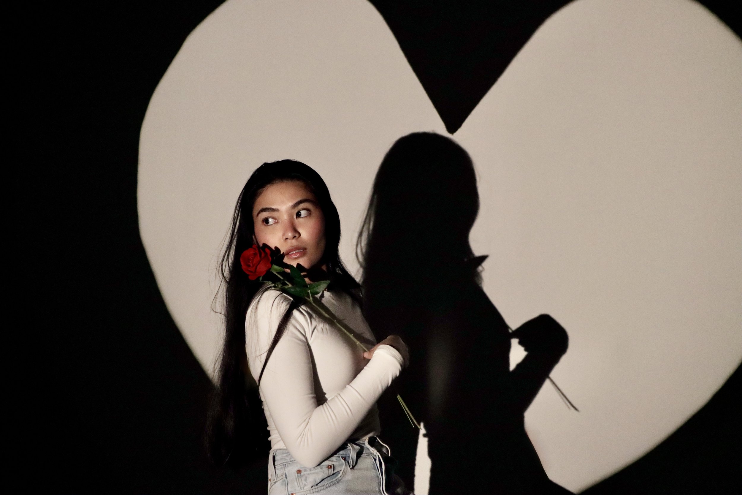 A young woman with long dark hair holding a red rose, standing in front of a black and white heart-shaped shadow background, with her shadow visible on the wall.