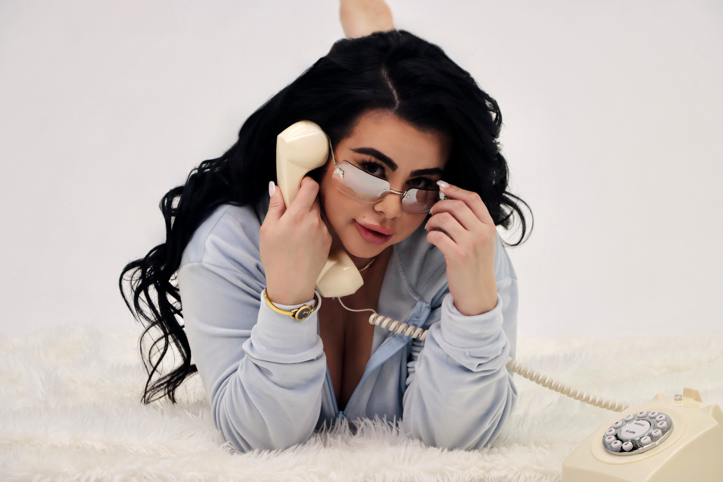 A woman with long dark hair, wearing sunglasses and gold jewelry, lies on a soft white surface while speaking on a vintage beige rotary phone.