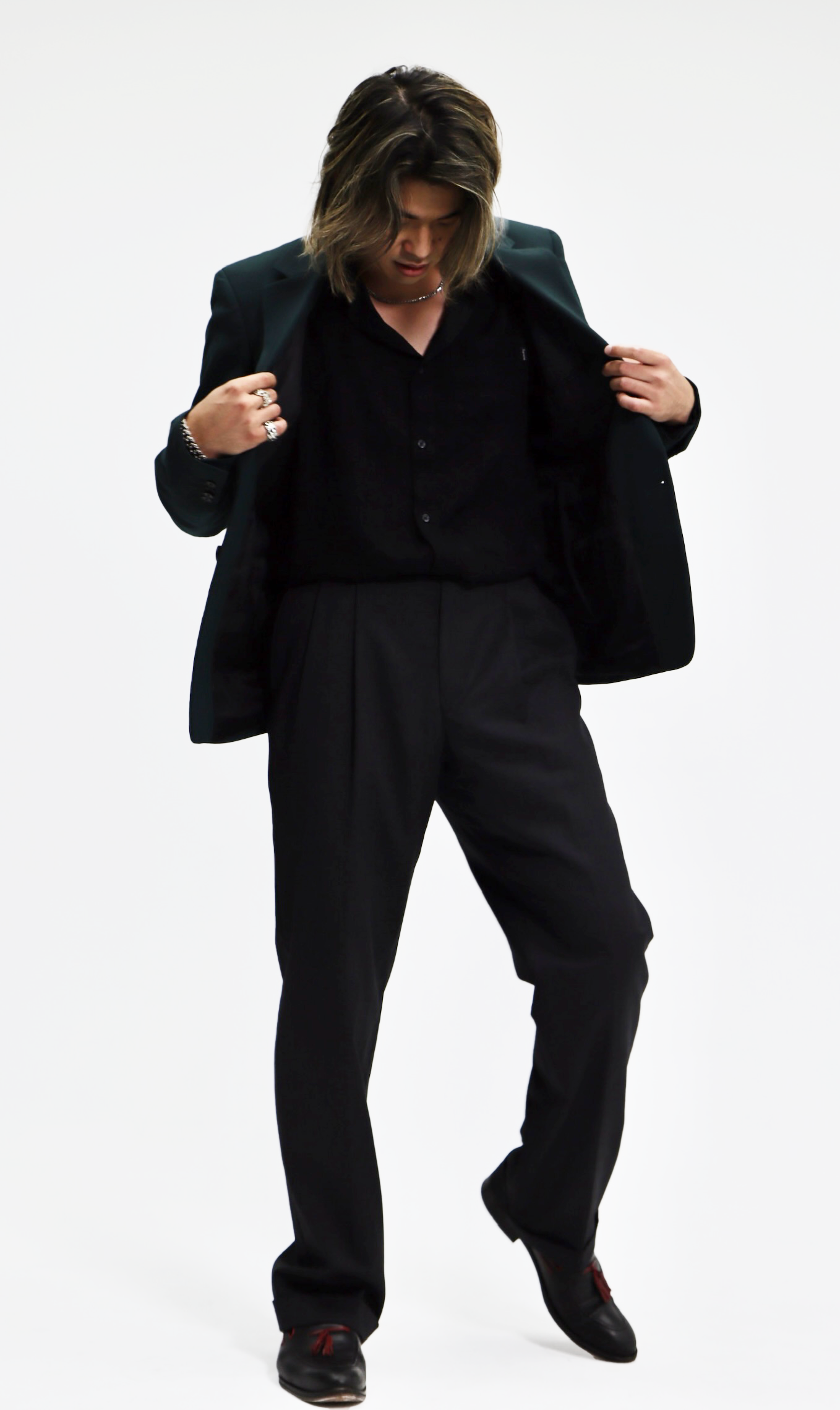 Person with long hair in a black suit, black shirt, and black shoes, standing against a white background.