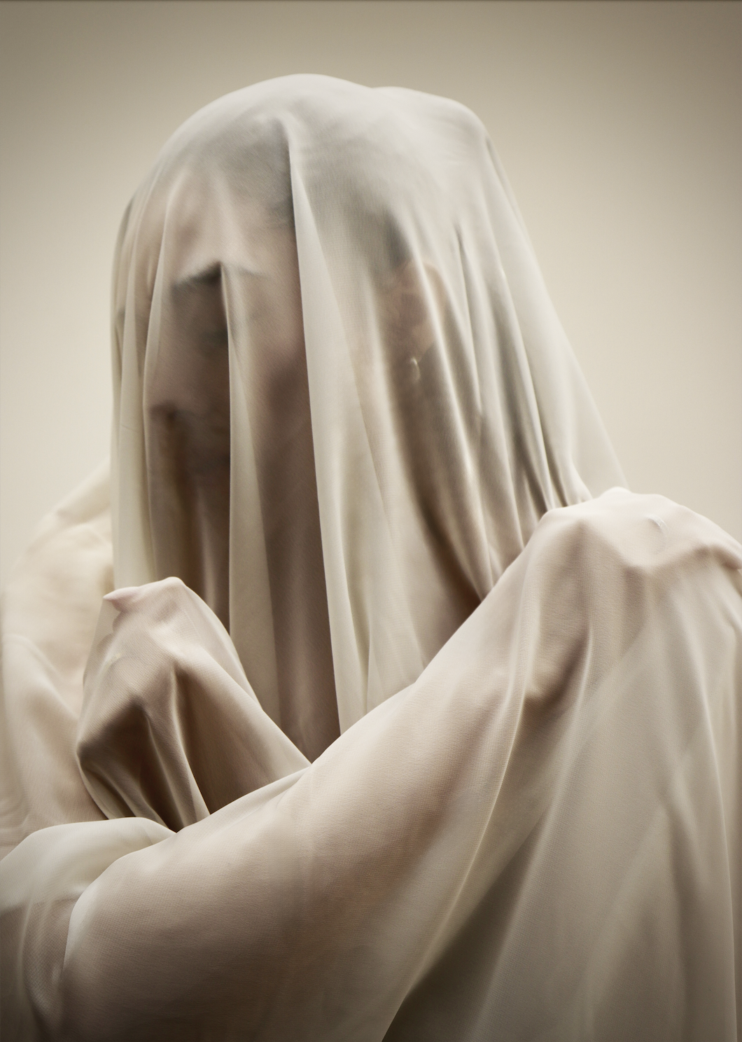 Person with head and shoulders covered by sheer, beige fabric, creating a ghostly appearance.