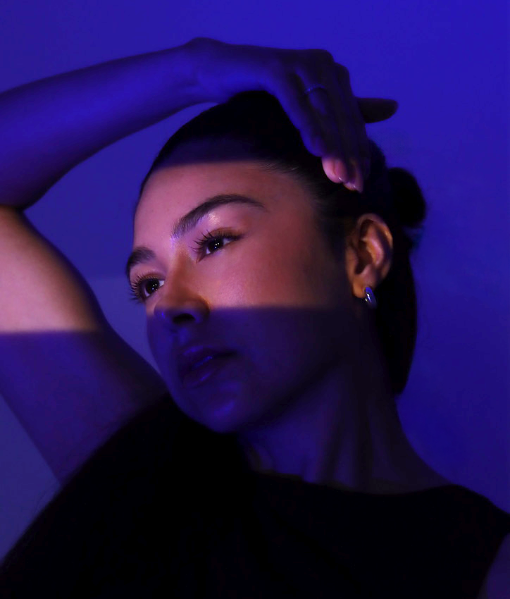 A woman with dark hair and an earring, illuminated by blue and purple lighting, with her hand resting on her head.