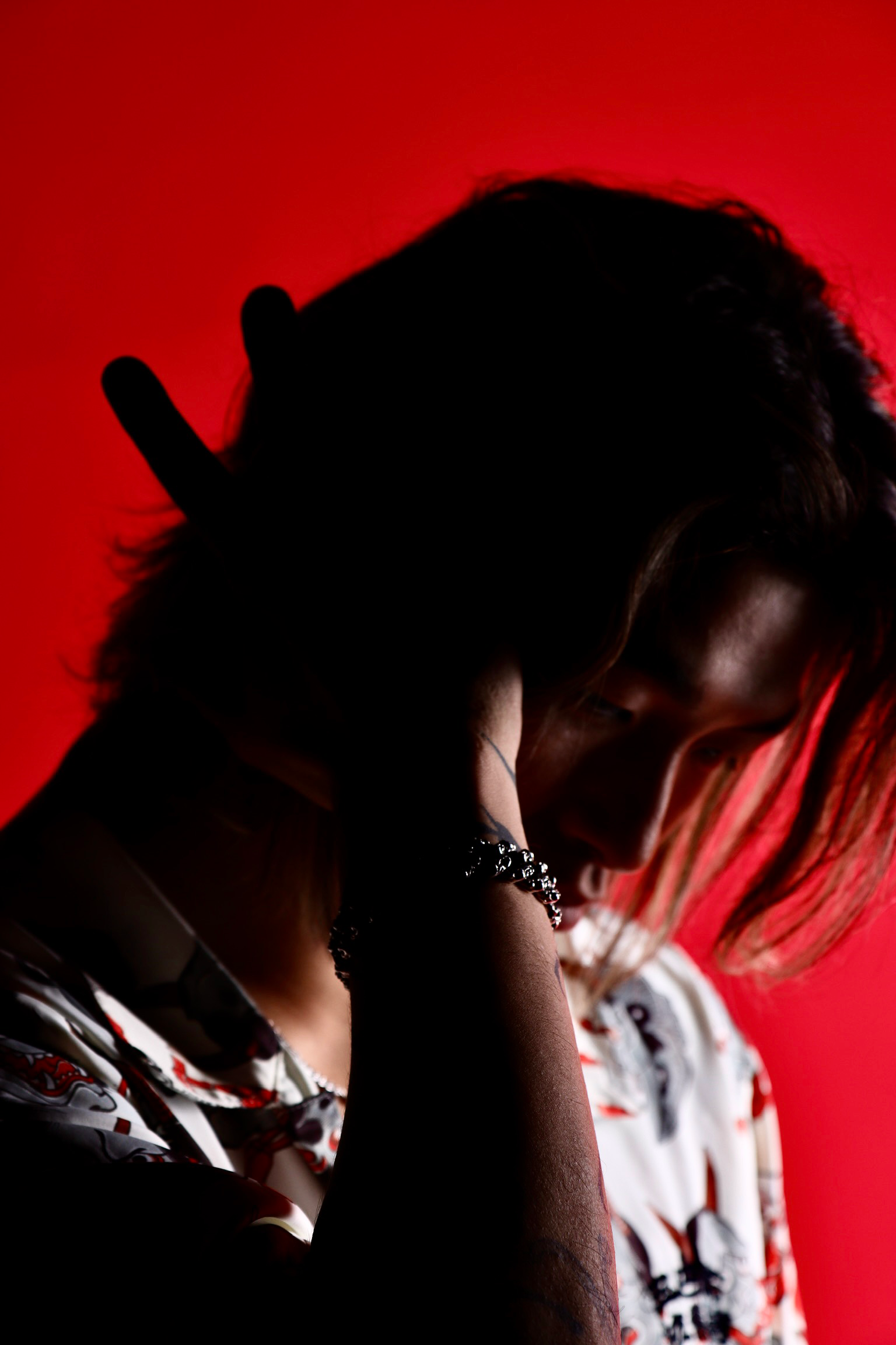 Silhouette of a person with long hair, wearing a bracelet, touching their face, against a red background, with a blurred background of another person.