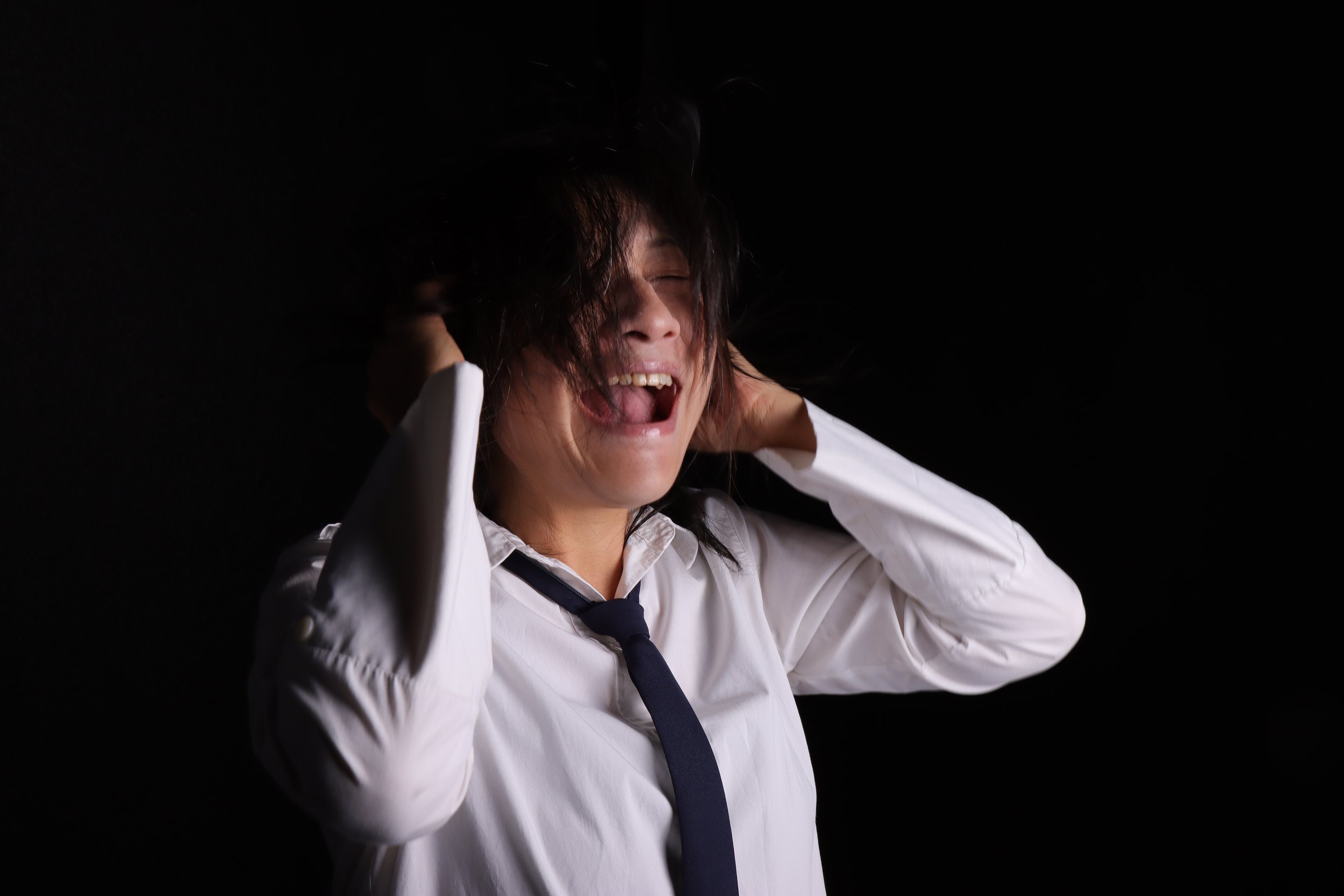 A woman in a white shirt and dark tie with long dark hair, holding her head with her hands, appears distressed or frustrated against a black background.