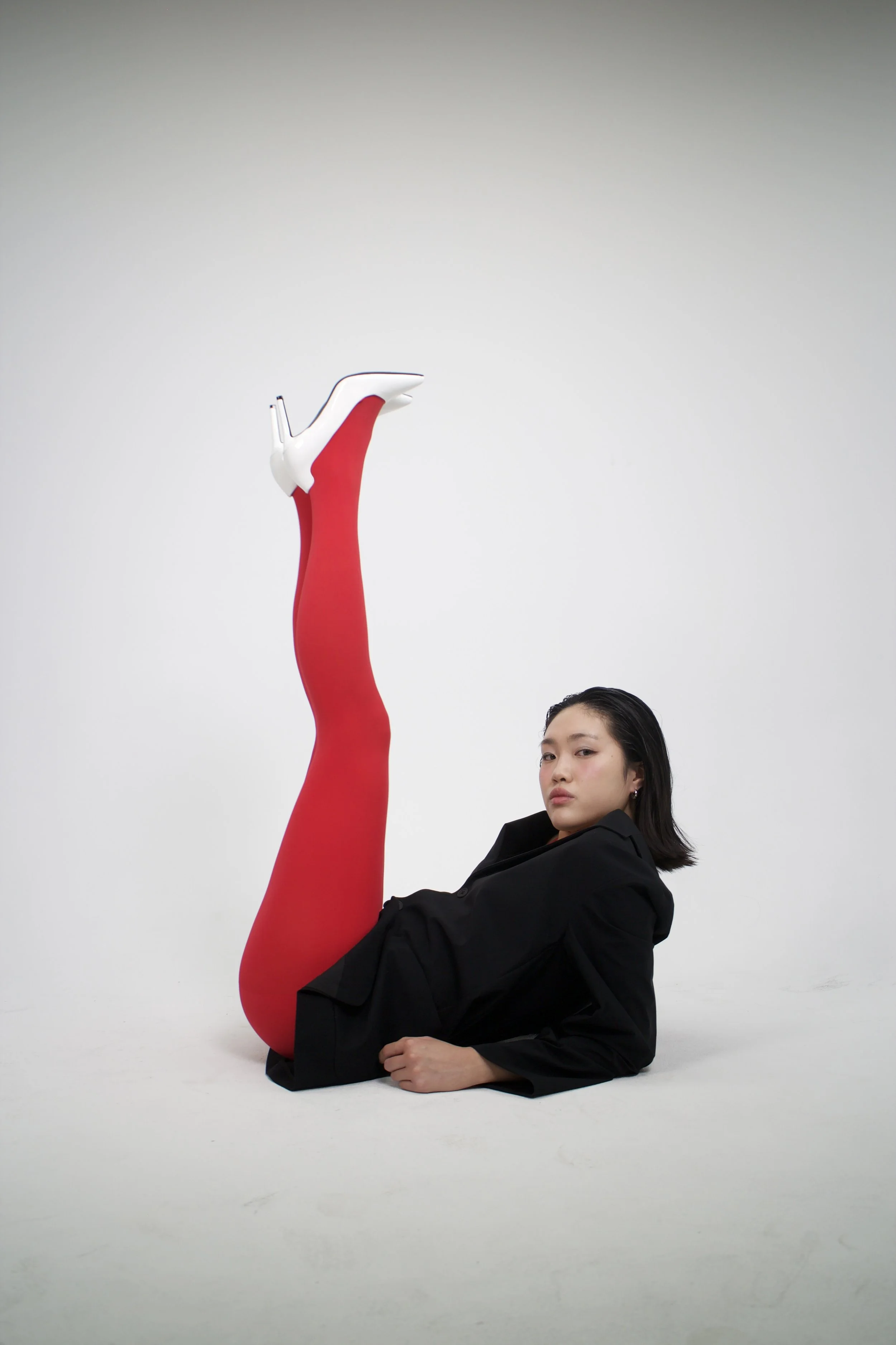 A woman lying on the floor with one leg raised straight up, wearing red tights and white high heels, against a plain white background.