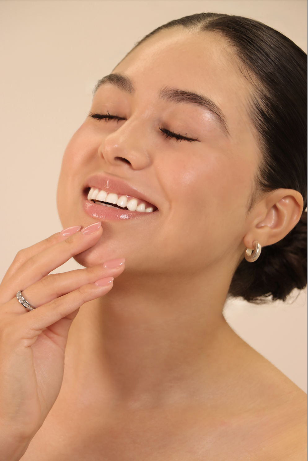 Close-up of a smiling woman with closed eyes, touching her chin with her hand, wearing earrings and a ring.