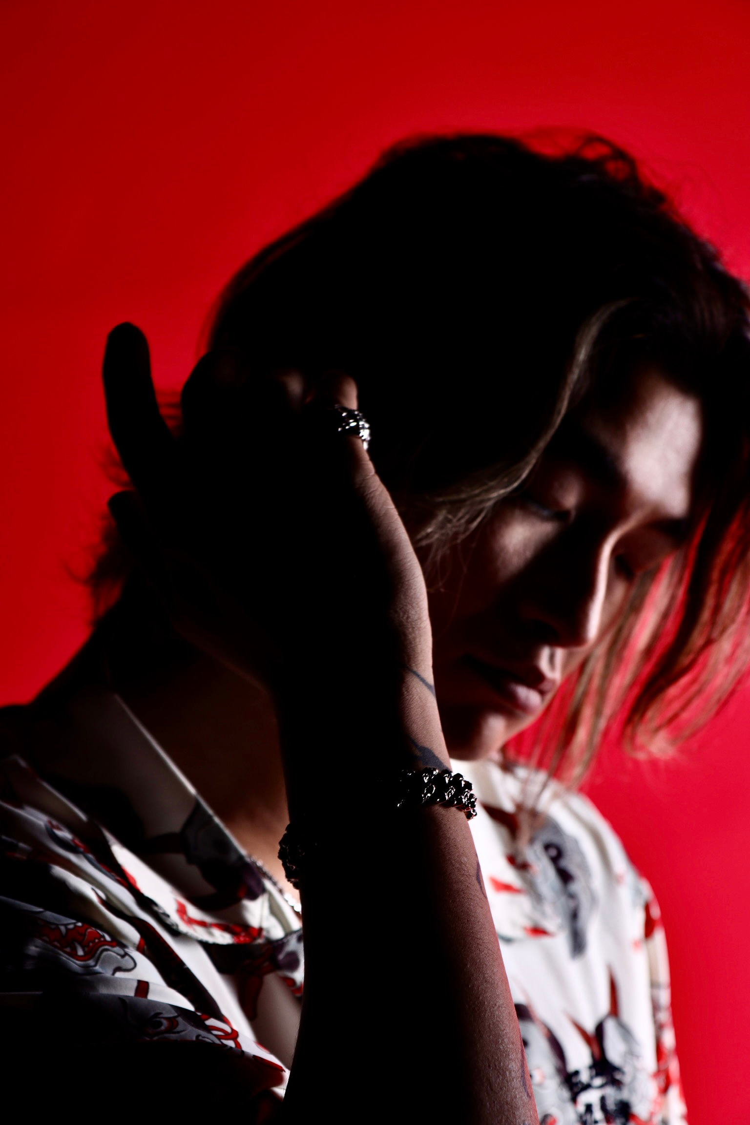 A person with medium skin tone and brown hair styled in loose waves, facing downward with eyes closed, holding their head with one hand. The background is solid red. The person is wearing rings, a bracelet, and a shirt with a colorful pattern.